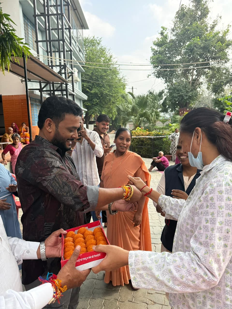 A smiling man has a rakhi tied on his wrist while a woman holds out a box of laddoos; people around them smile