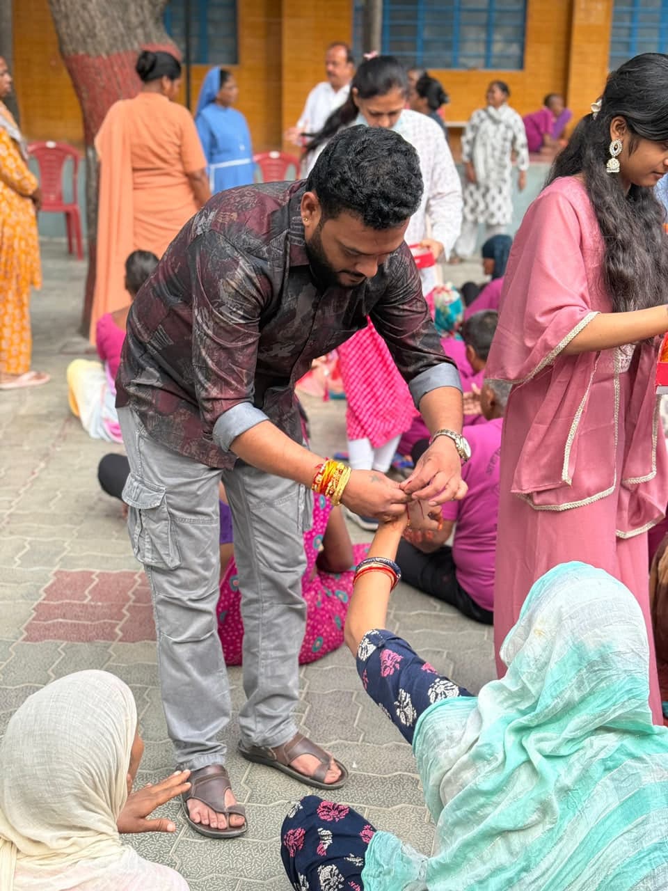 A man bends to tie a rakhi thread on the raised wrist of a seated woman, among other residents seated in a courtyard