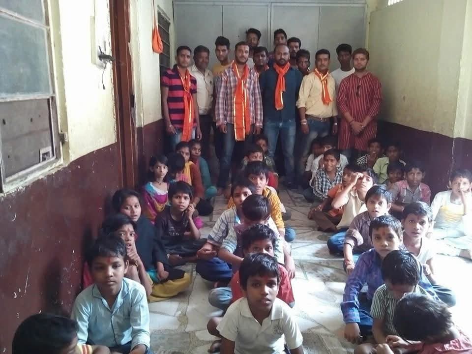 Volunteers wearing saffron scarves stand behind rows of children seated on the floor of a corridor