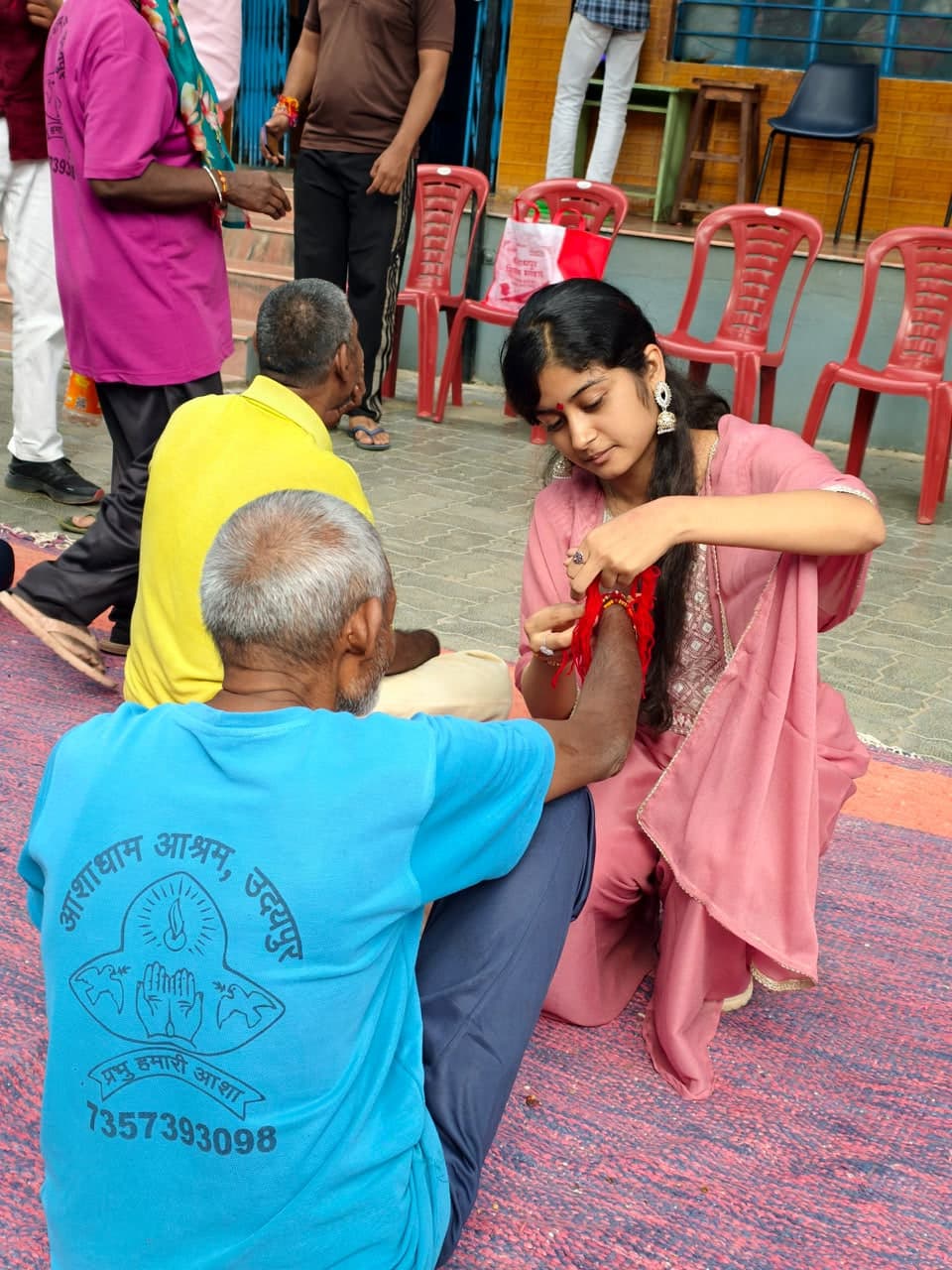 A young woman kneels to tie a red rakhi thread on the wrist of an elderly man seated on a mat