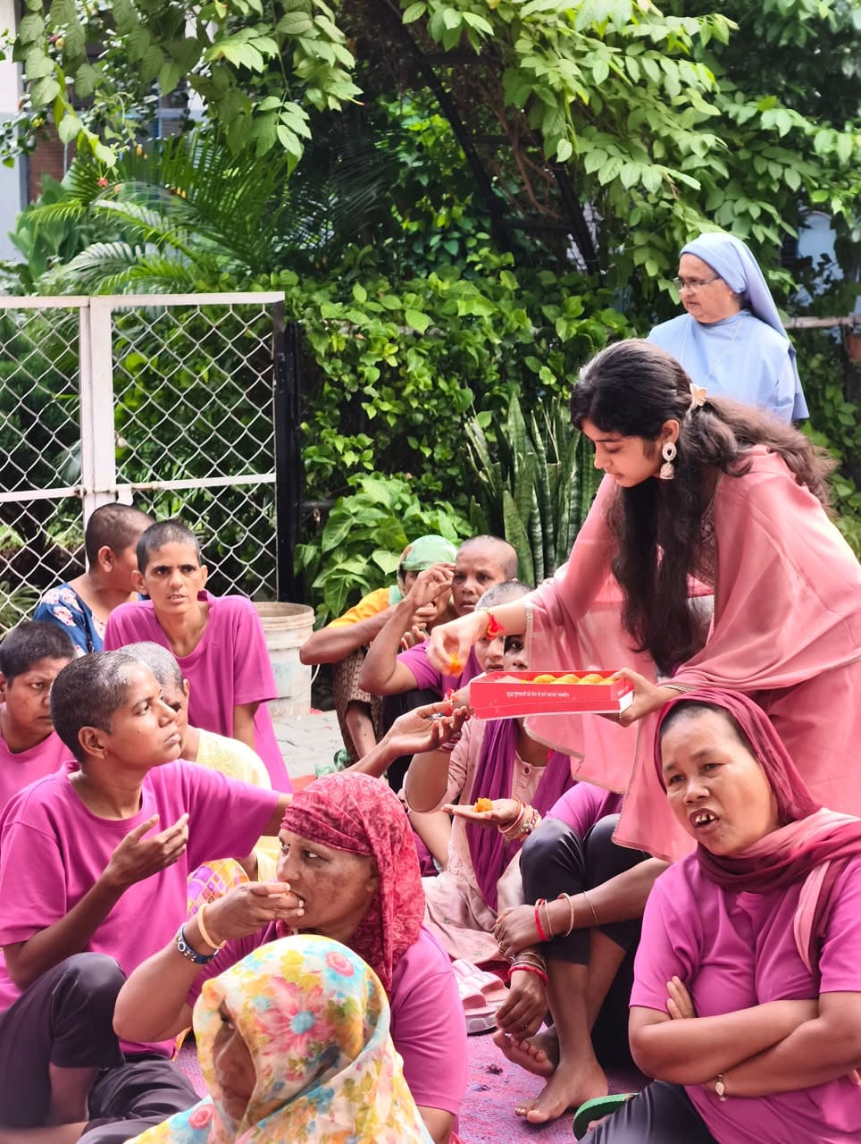 A young woman in a pink suit offers sweets from a box to women seated on the ground, with trees and a nun in the background